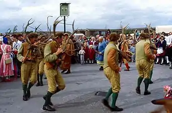 Photo en couleurs d'hommes vêtus d'habits verts, jaunes et marron qui dansent en rond en portant dans les mains des bois de renne