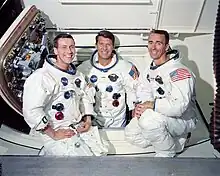 Photographie en couleur des trois membres d'équipages d'Apollo 7 souriants en combinaison blanches devant le module de commande Apollo.