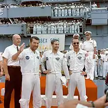 Trois hommes en salopette blanche de la NASA. Nombreux marins à l'arrière-plan.