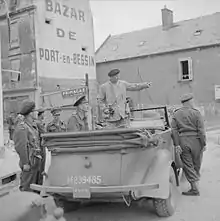 Photographie montrant un véhicule décapoté occupé par deux personnalités dont une debout et entouré de cinq personnes.