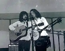 Photo en noir et blanc de deux hommes en train de jouer de la guitare et de chanter dans le même micro