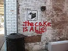Photo du mur d'une ruelle, sur lequel sont graffés la phrase "The cake is a lie!" en rouge, et l'icône de gâteau de Portal