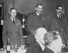 Trois hommes en uniforme militaire devant une table de restaurant où sont assis d'autres convives.