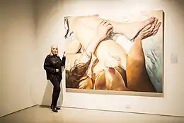 Une femme âgée à côté d'un de ses grands tableaux, représentant une femme nue