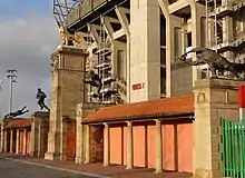 Photographie en couleurs. Aux abords d'un stade qui s'élève sur la droite, une porte est surmontée de sculptures d'un lion et de joueurs de rugby.