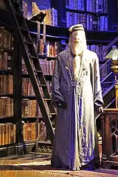 Un pantin debout vêtu d'une longue robe masculine grise, placé devant une grande bibliothèque de livres anciens sur un mur circulaire.