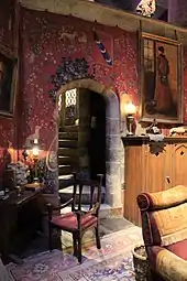 Une salle chaleureuse aux teintes rouges, avec tapisseries médiévales, tapis, fauteuil, meubles en bois et escalier de pierre en colimaçon menant à l'étage.
