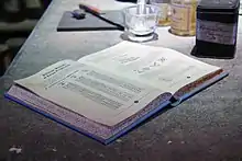 Photo d'un livre ouvert posé sur une table en bois. Le livre à la couverture bleue et aux tranches mouchetées présente des colonnes de symboles. À l'arrière-plan se trouve une planche à découper avec un couteau, et quelques bocaux.