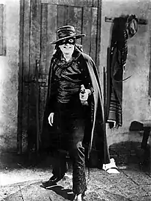 Photographie en noir et blanc d'un homme portant un chapeau, un masque et une cape noire, tenant dans sa main gauche une épée.