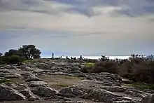 Au premier plan de la photo, un plateau rocheux avec de la végétation sur les côtés, des gens sont dans le fond. Au second plan, la mer, les montagnes au loin et le ciel dominent le reste de la photo.