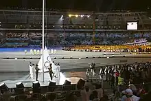 Au premie plan, le drapeau olympique en phase d'être gissé sur un mat. Dans le fond, ds athlètes devant la tribune opposée.