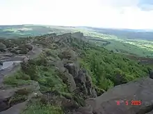 au premier plan, le sommet d'une falaise composée de vastes blocs gris entrecoupés de buissons, dominant un paysage de bocage qui s'étend en arrière plan