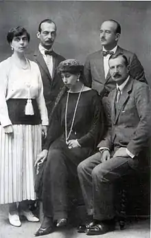 Photographie en noir et blanc montrant deux femmes portant des robes et trois hommes en costumes.
