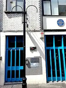 Photo de l'entrée d'un immeuble aux murs blancs, fermée par de grandes grilles bleues.