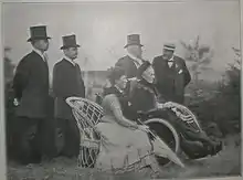 Une dame assise sur un fauteuil en rotin et tenant une ombrelle fermée est assise à côté de Marie-Henriette dans un fauteuil roulant, vêtue d'une robe sombre et enveloppée dans une couverture, dont le visage de profil est coiffé d'un petit chapeau sombre, derrière les dames, se tiennent debout quatre messieurs en costume civil et portant un chapeau haut-de-forme, hormis le dernier qui arbore un canotier