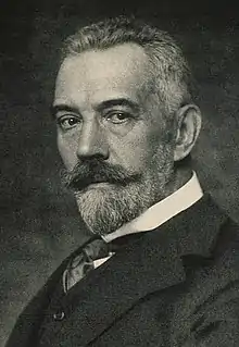 portrait photographié en noir et blanc d'un homme en costume aux cheveux blancs courts et portant barbe et moustache