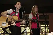 Deux jeunes filles en train de chanter (une brune à gauche jouant de la guitare et une blonde à droite). Elles portent des gilets rose foncé sans manches sur des chemisiers blancs, avec des cravates vertes visibles