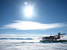Avion à hélices posé sur un immense glacier près de barils verts, avec des montagnes en fond, sous un soleil éblouissant.