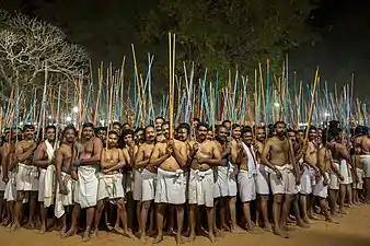 Une troupe d'hommes, de face, vêtus d'un pagne blanc, pieds nus, brandissent de grand bâtons.