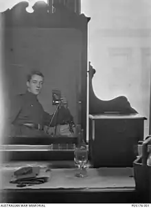 Une commode avec un grand miroir et une bouteille de vin, un verre et quelques papiers posés dessus. Un homme en uniforme militaire est visible dans le reflet du miroir. Il a la main dans sa poche et utilise un appareil photo sur un trépied.