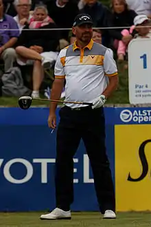 Homme en pied avec une casquette, de face, tenant un club de golf dans la main gauche.