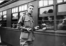 Un jeune homme en uniforme militaire tenant quelque chose dans ses mains se tient à côté d'un wagon de train où des hommes regardent à l'extérieur.