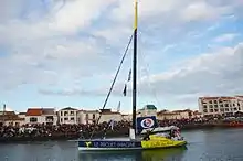 Un voilier bleu marine et jaune descend un chenal au moteur. Sur la coque, un colibri et les inscriptions « Le Projet Imagine » et « Le Souffle du Nord ». Une foule est massée sur le quai.