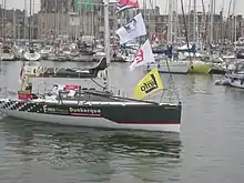 Un voilier noir et blanc avance au moteur dans un port. Sur la coque, on peut lire « Faber France » et « Dunkerque communauté urbaine ».