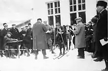 Photographie en noir et blanc d'homme sur des skis, tenant un bâton dans chaque main et portant le dossard numéro 81,s'apprêtant à prendre le départ, entouré de plusieurs hommes dont un skieur avec le dossard numéro 82 et deux hommes assis à une table sur sa gauche.