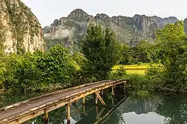 Vue de trois-quarts d'une passerelle en bois au-dessus d'un lagon, arbres et montagnes karstiques, tôt matin dans les rizières de Vang Vieng.