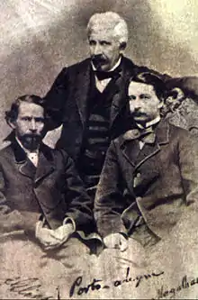 Une vieille photo représentant deux hommes aux cheveux noirs assis au premier plan et un homme debout à cheveux blancs derrière