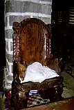 Photographie du trône royal d'Uvea se trouvant dans le palais. Le siège est fait en bois et on y trouve dessus un coussin.