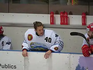 Photographie couleur d’un homme, équipé d’un équipement de hockeyeur sans casque, appuyé contre la bande depuis son banc