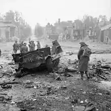 photo montrant des soldats passant à proximité d'un véhicule militaire détruit au milieu des ruines d'un village