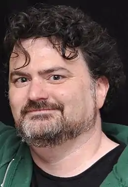 Photographie du visage d'un homme barbu portant un tee-shirt noir et un sweat vert