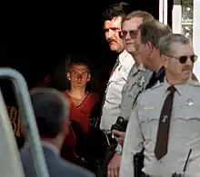 À droite, quatre policiers alignés en position d'attente alors qu'au fond un homme habillé d'une combinaison orange sort de l'ombre en marchant.
