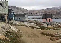 Photographie en couleurs de maisons en bois, l'une rose, une deuxième bleu foncé, une troisième gris foncé, bordant une route, les ruines d'une habitation au premier plan, les eaux d'un fjord visibles au second plan.