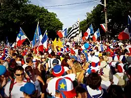 Photographie montrant le Tintamarre de Caraquet, lors du festival acadien. Les gens sont maquillés et habillés des couleurs du drapeau de l'Acadie et plusieurs personnes portent aussi des drapeaux.