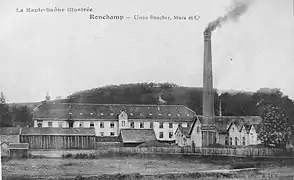 Photo noir et blanc montrant un long bâtiment industriel anciens surmonté d'une cheminée fumante et une petite colline proche.