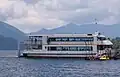 Photo couleur d'un bateau blanc à deux étages sur une étendue d'eau, une chaîne de montagnes et un ciel nuageux en arrière-plan.