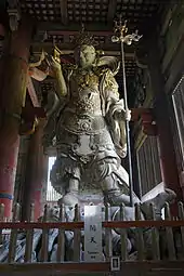 Sculpture en bois vue en contre-plongée d’un dieu en armes et en armure, air féroce.