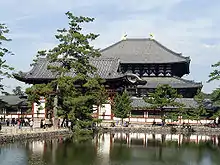Photo couleur d'un imposant édifice religieux bouddhique en bois, à deux étages. Arrangement de poutres et de panneaux blancs, avec deux toitures de tuiles grises, sur fond de ciel bleu. Au premier plan, quelques dizaines de personnes se tiennent debout, dans l'allée qui mène aux marches de l'entrée du bâtiment.