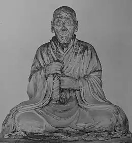 Statue assise d’un vieil homme égrainant un chapelet. Noir et blanc.