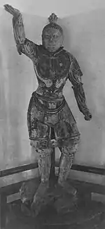 Tamonten, un des Quatre Rois célestes in the Kaidan Hall. Portrait of a statue in front view. The right arm is raised, the hair sculpted with a top knot and the breast with armour. Narrow slit eyes and a facial expression as if frowning.