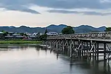 Dans une atmosphère grise lumineuse un pont avec de nombreux piliers de bois enjambe une belle rivière. Il arrive à une petite ville blottie entre montagne, campagne.