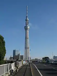 Photo couleur d'une tour métallique surmontée d'une antenne de daiodiffusion, sous un ciel bleu. Un trottoir et une route à quatre voies au premier plan.