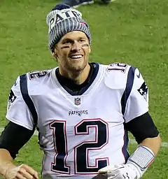 Portrait de Tom Brady, souriant avec les poings fermés et un bonnet sur la tête.