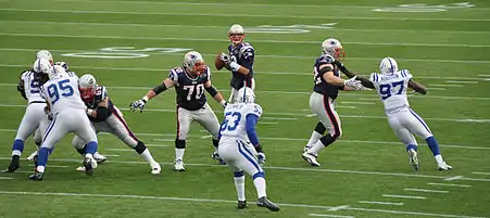 Tom Brady, en maillot bleu avec le ballon en main, protégé par sa ligne offensive sur un terrain de football américain.