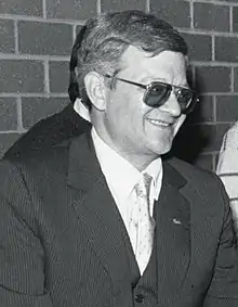  Un homme cinquantenaire, lunette de soleil et costume trois pièces et cravate.