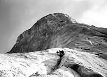 Photographie en noir et blanc montrant une personne escaladant une falaise, vue depuis le sol